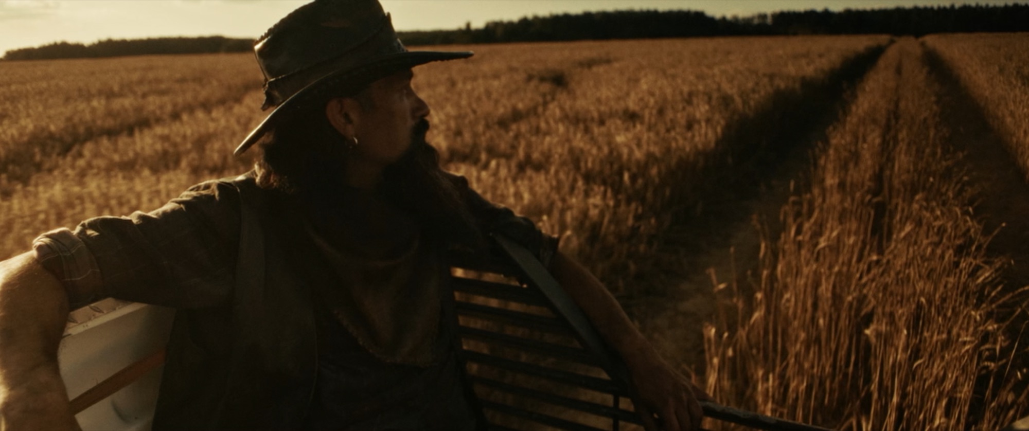 Bearded man in a cowboy hat riding on a moving vehicle beside a golden wheat field at sunset - Amon Amarth